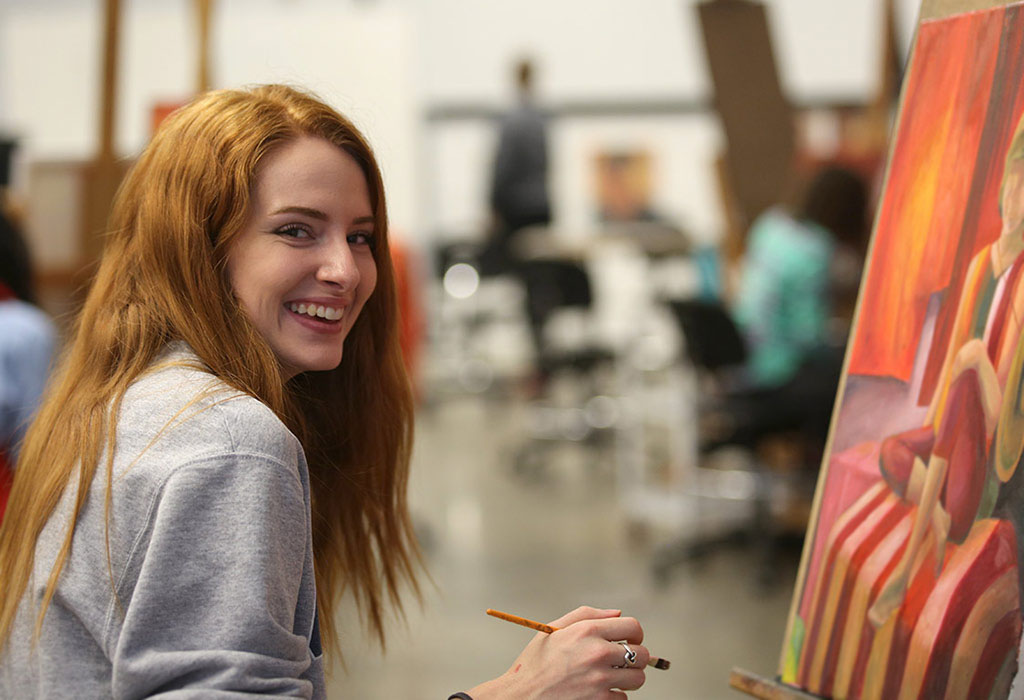 A student seated at a painting with brush in hand. They've paused their work to smile at the camera. 