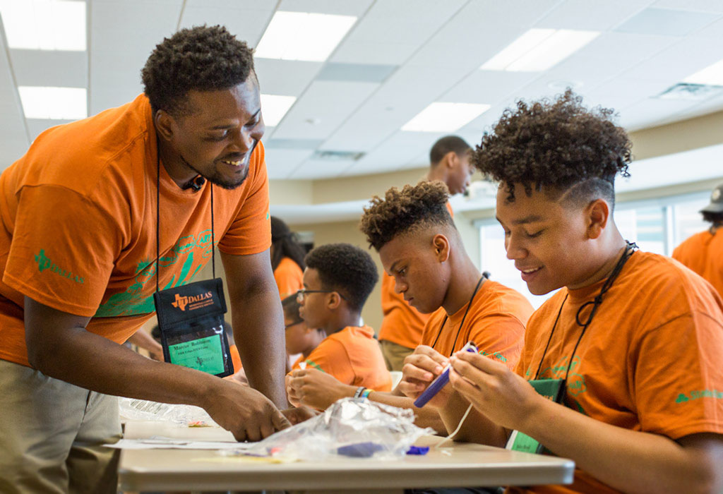 A counselor and several students, all wearing orange t-shirts chat over a table at a camp event. The students are working on a craft with their hands. 